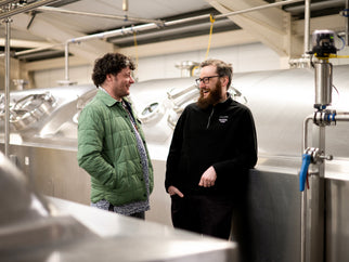 Two men talking in a brewing facility with large equipment around them.